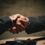 木製テーブルを挟んで二人が握手する様子。信頼と誠実な約束を象徴するイメージ
